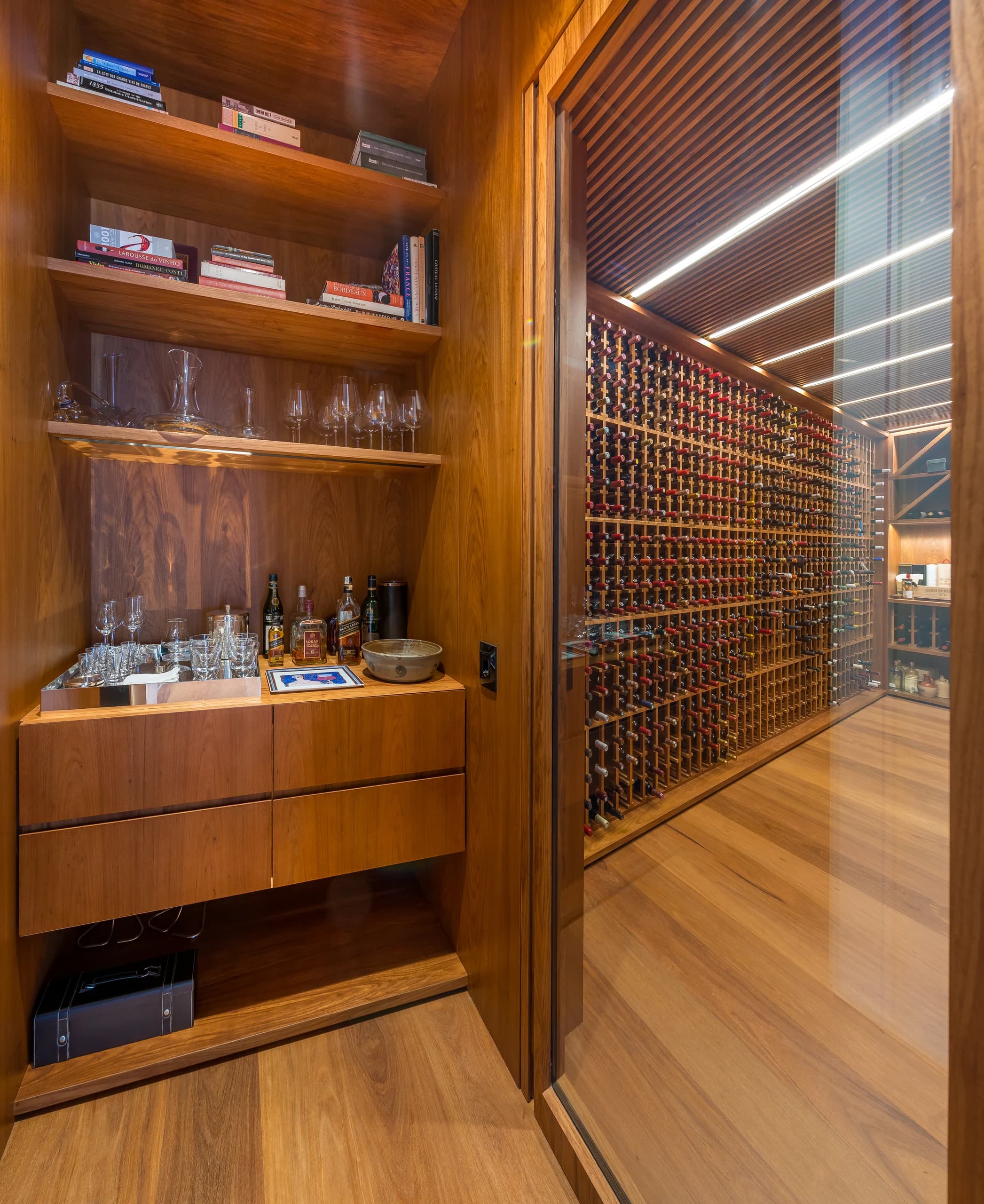 Vista vertical da Adega Andiamo através do fechamento envidraçado, junto a um móvel externo com livros, taças e bebidas; escaninhos e forro ripado visíveis no interior.