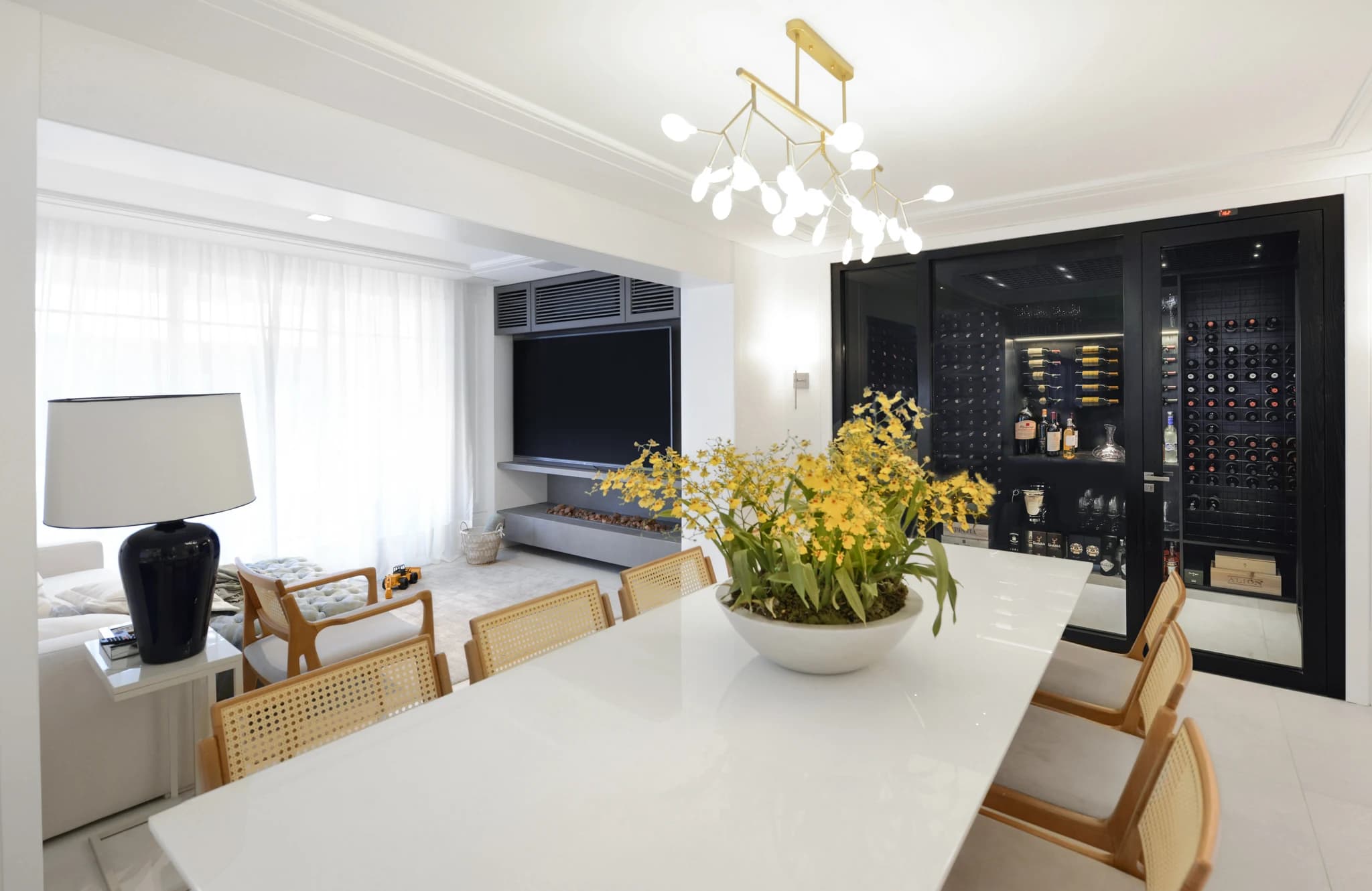 Vista ampla da sala com mesa de jantar, arranjo de flores amarelas e a adega General Mena Barreto ao fundo, à direita.